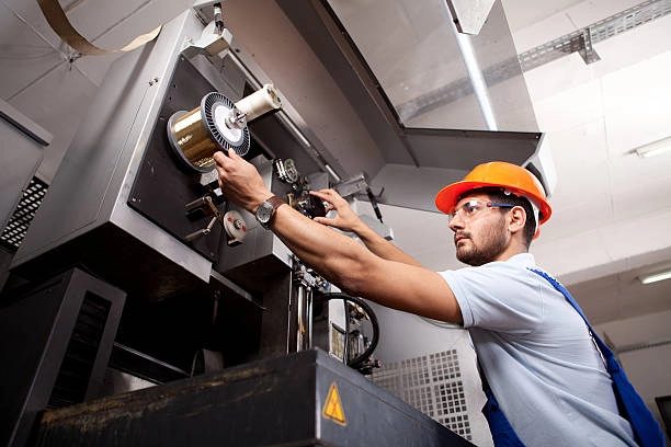 The image shows a factory worker wearing a safety helmet and goggles operating a large industrial machine