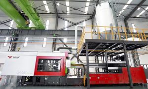 The image shows a large industrial injection molding machine installed in a modern factory with overhead cranes and material silos